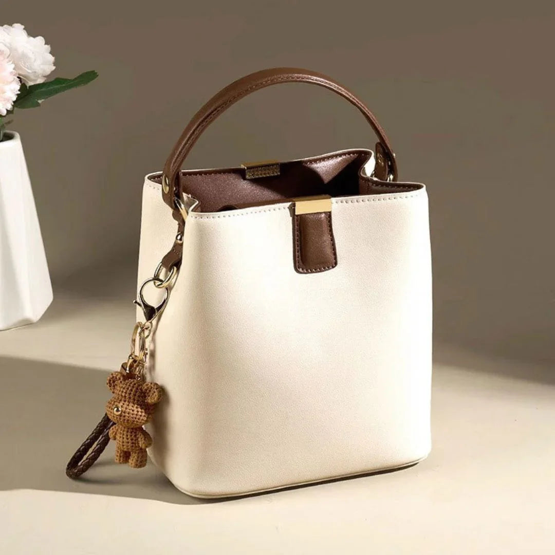 Cream and brown bucket handbag with bear keychain on beige surface, vase with flowers nearby