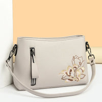 Beige leather crossbody bag with floral embroidery and front zipper, against pastel background.