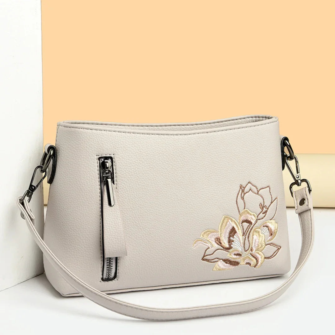Beige leather crossbody bag with floral embroidery and front zipper, against pastel background.