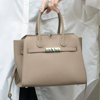 Taupe leather handbag with top handles and silver hardware, held by person in white shirt