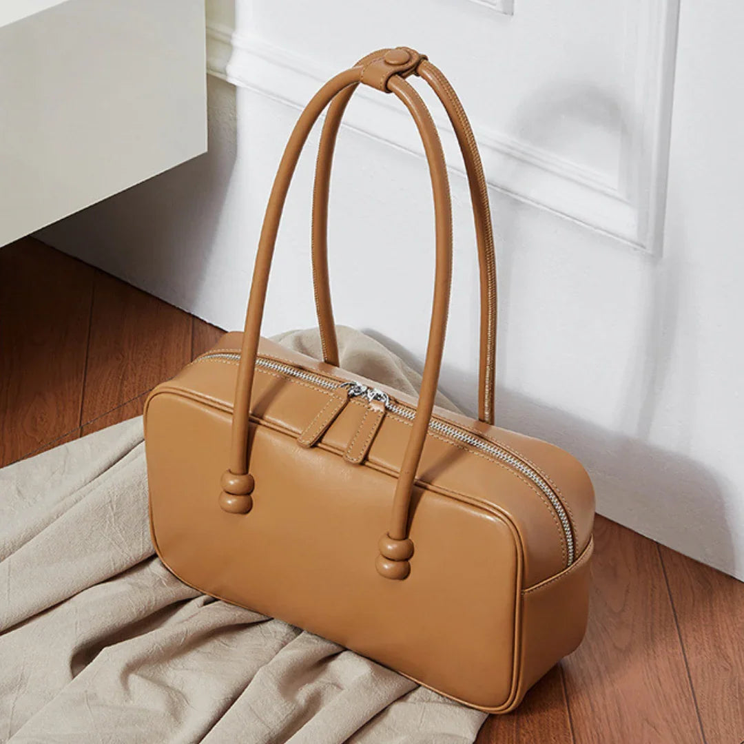 Tan leather handbag with double handles on wooden floor near white wall