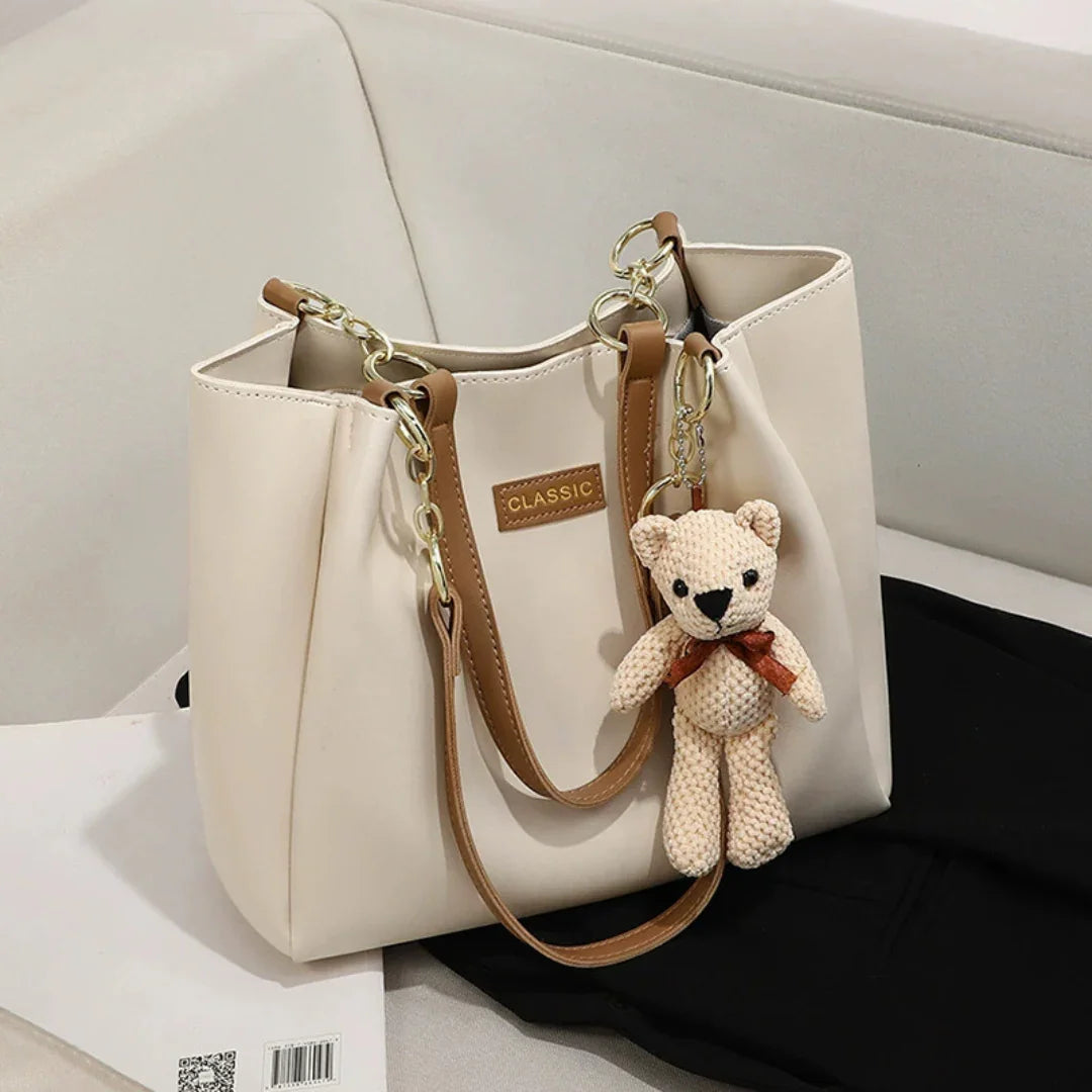 Beige tote bag with brown handles and teddy bear keychain on white sofa