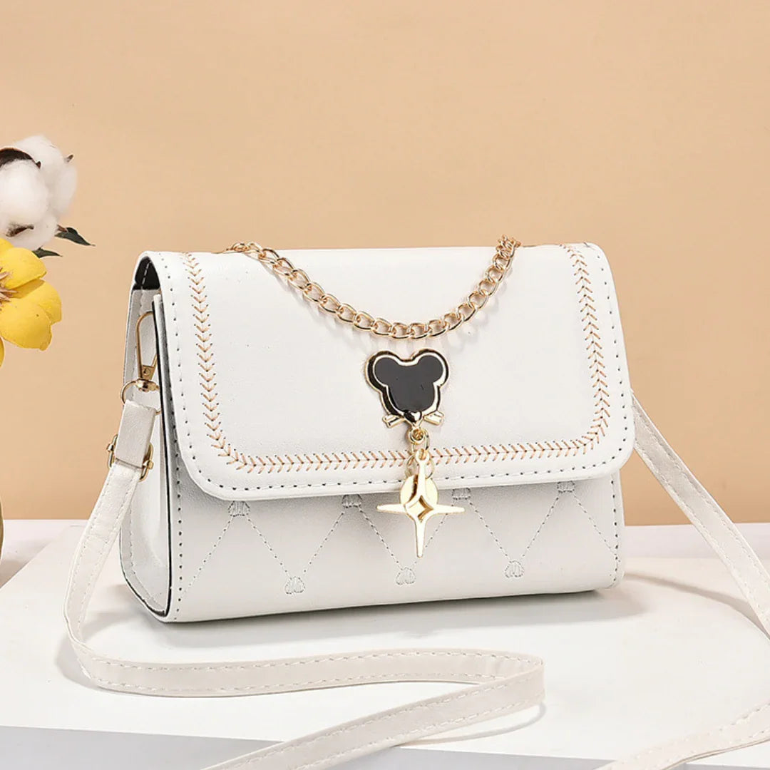 White crossbody bag with gold chain and black bear pendant, on beige background