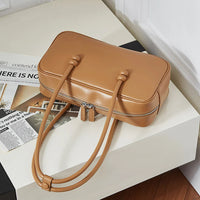 Tan leather rectangular handbag with double handles on white table beside magazines