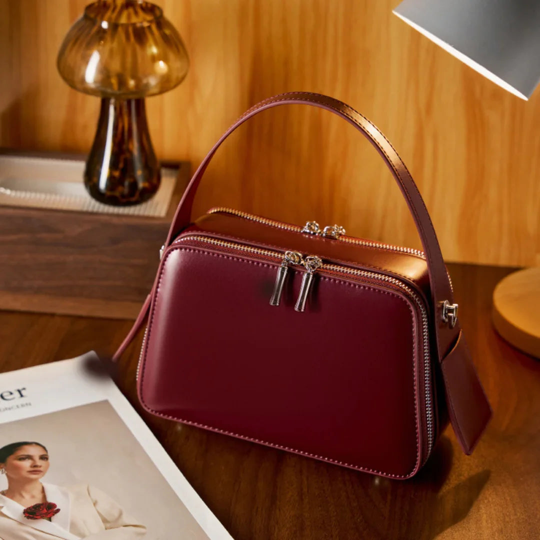 Burgundy leather handbag with top handle on wooden desk, next to lamp and magazine
