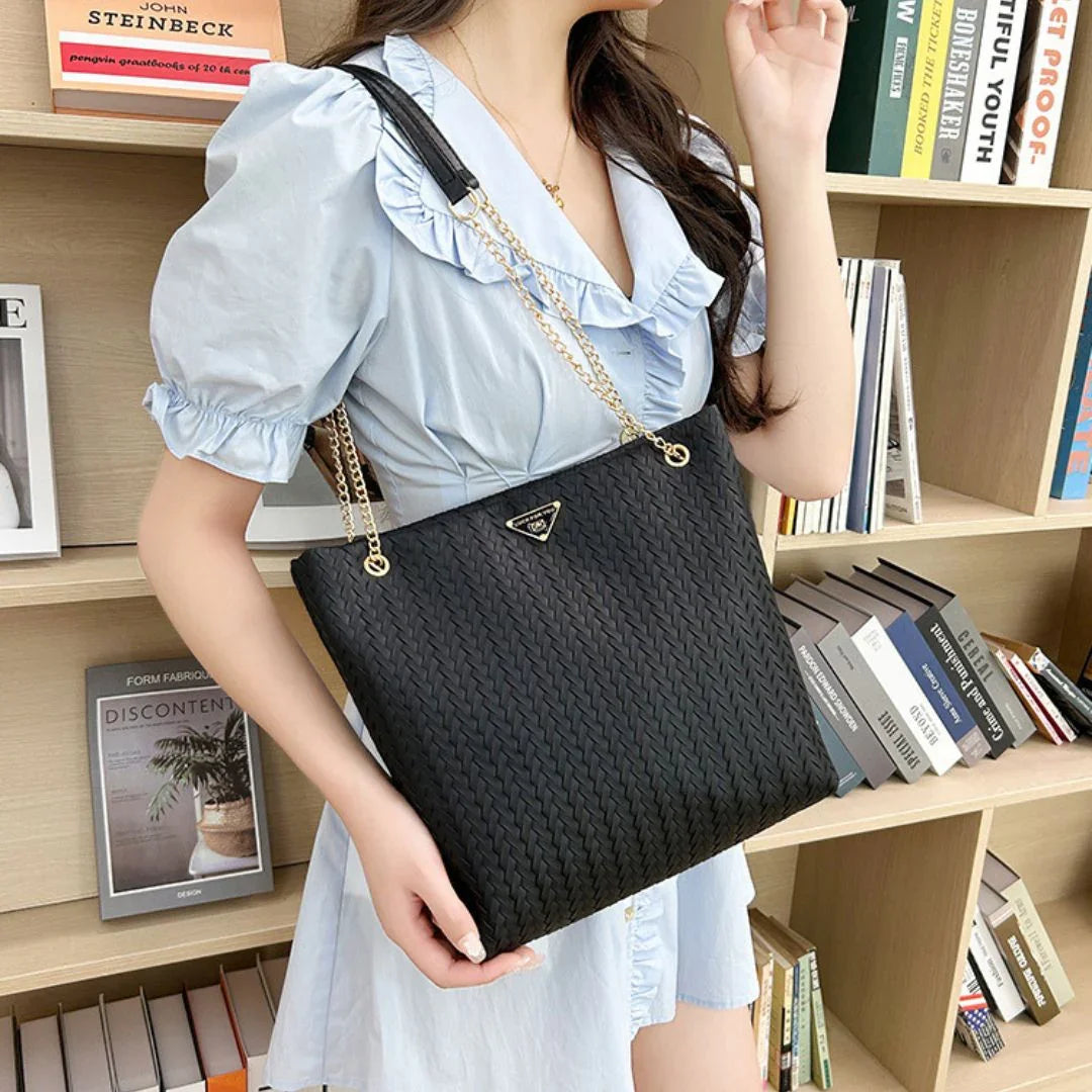 Woman in light blue dress carrying black woven tote bag with chain strap in front of bookshelf