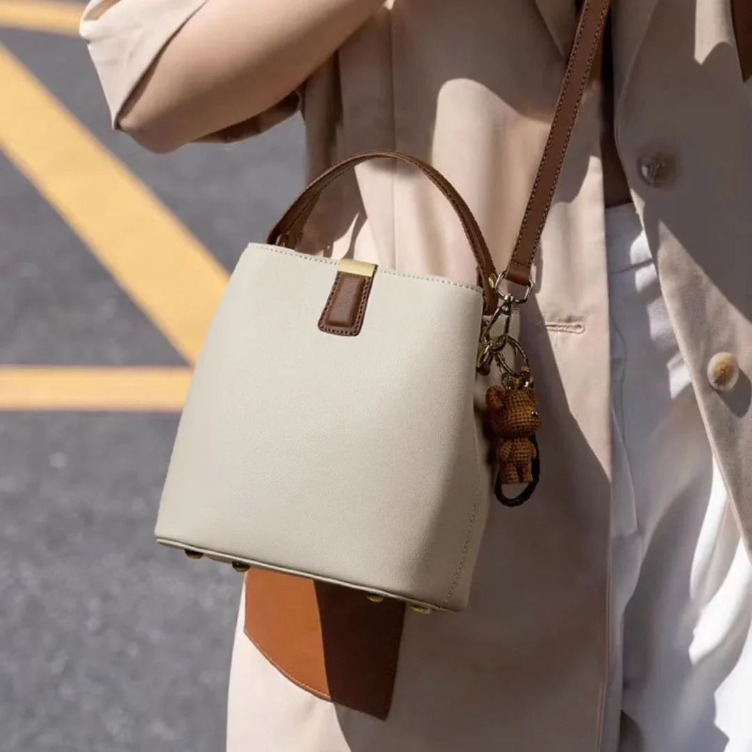 Beige leather handbag with brown strap and teddy bear keychain, worn over trench coat outdoors