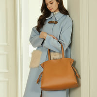 Woman in light blue coat with faux fur pockets holding a large tan leather tote bag indoors