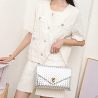 Woman in textured white outfit holding a silver woven clutch handbag indoors