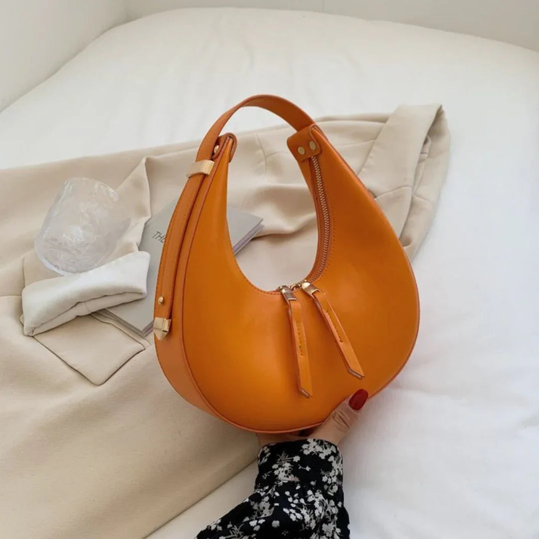 Orange crescent leather handbag on beige bedding, stylish purse with gold zipper accent