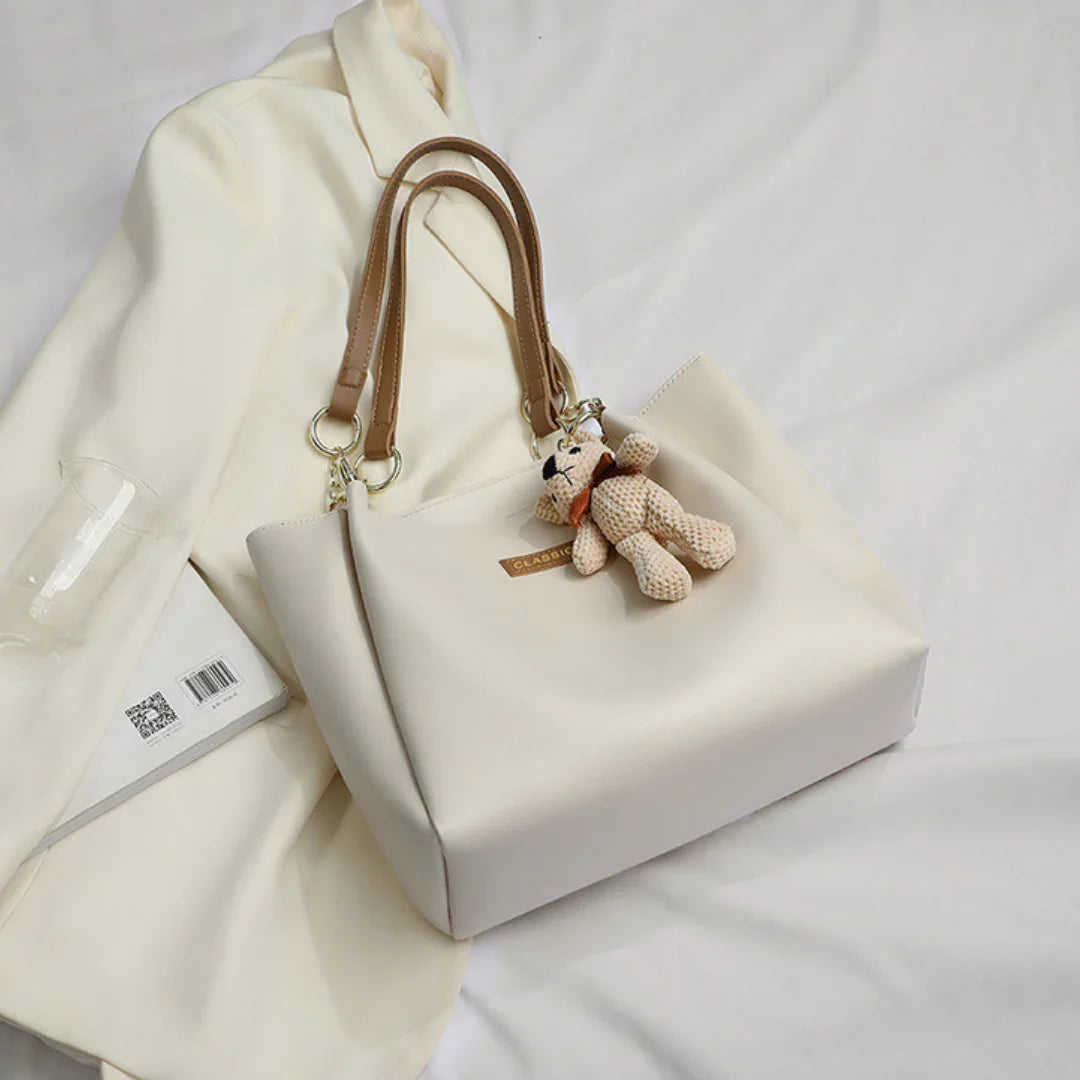 Cream handbag with brown handles and teddy bear keychain on white blazer and book
