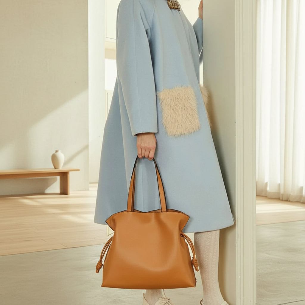 Woman in light blue coat with fur pocket holding tan leather tote bag in minimal room