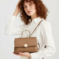 Woman in white blouse holding tan leather crossbody handbag with gold clasp