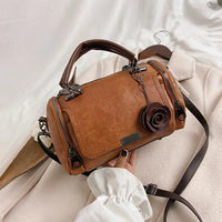Brown leather handbag with rose charm, top handle, and crossbody strap on beige coat