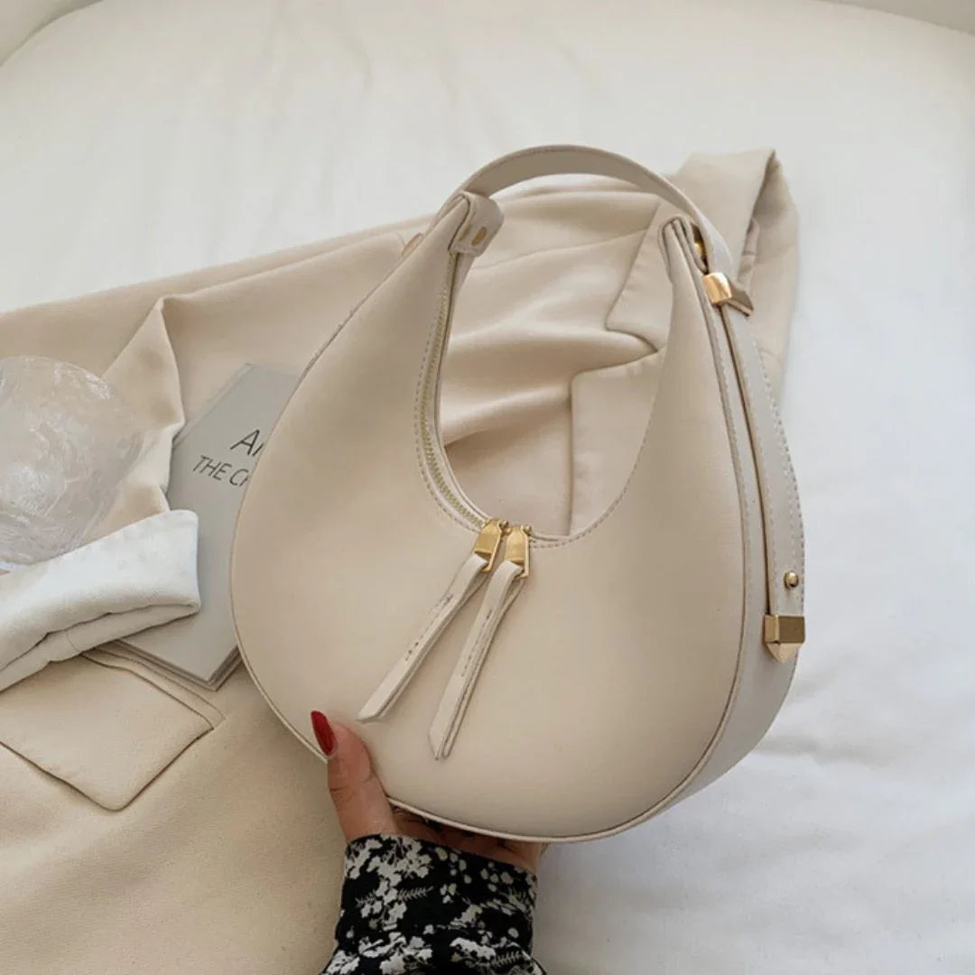 Beige crescent shoulder bag with gold zipper on a soft cream fabric background