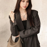 Woman in dark gray blazer with taupe leather handbag and silver necklace, neutral background