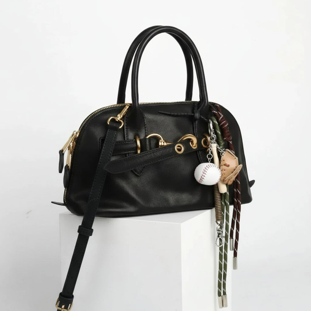 Black leather handbag with gold hardware and baseball-themed keychain, white background