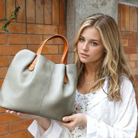 Woman holding large gray leather tote bag with brown handles, outdoor urban background