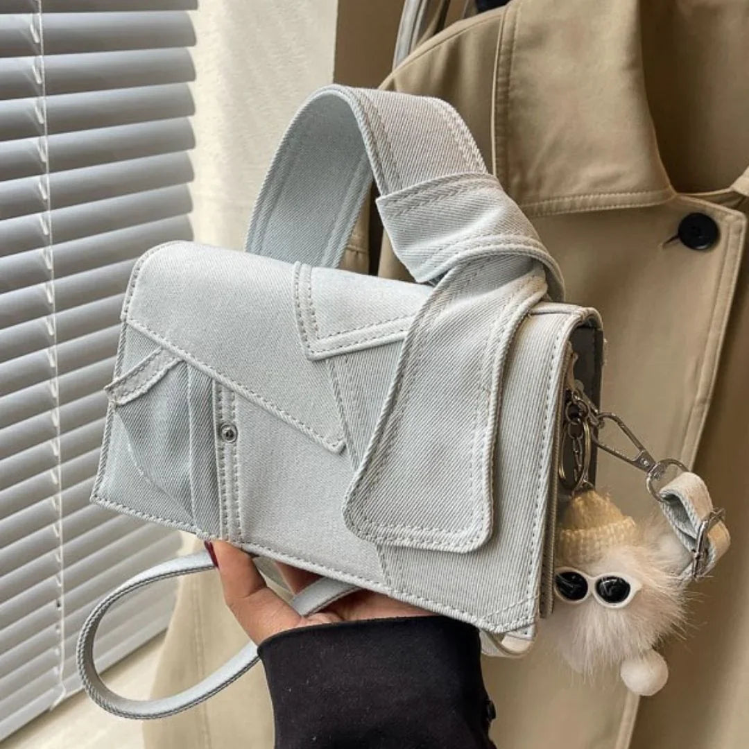 Light grey denim shoulder bag with wide strap and plush keychain, held by a window