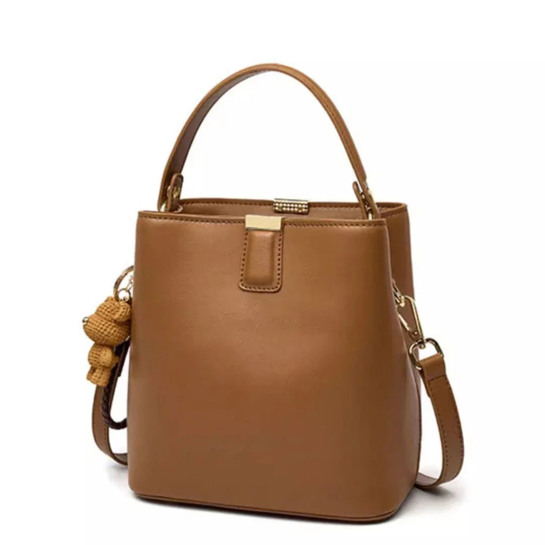 Tan leather bucket bag with top handle, crossbody strap, and woven bear keychain