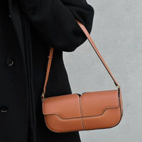 Brown leather shoulder bag with white stitching, worn with a black coat against gray background