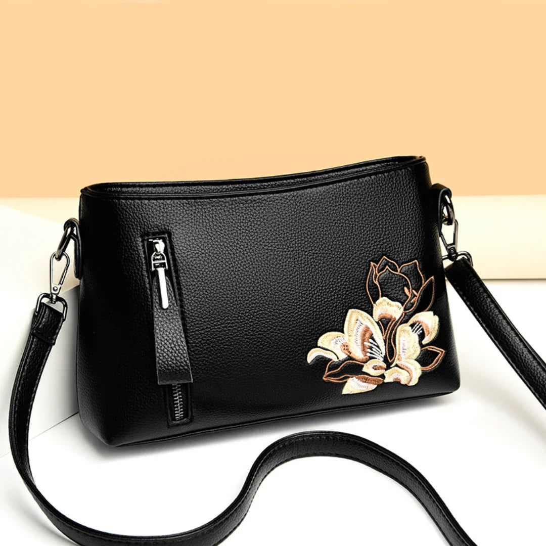 Black leather crossbody bag with floral embroidery and zip detail on a beige background
