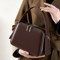 Woman holding brown leather box handbag with silver zippers, wearing brown trench coat