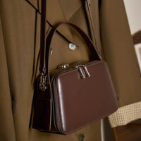 Brown leather crossbody box bag with silver zippers on a tan trench coat