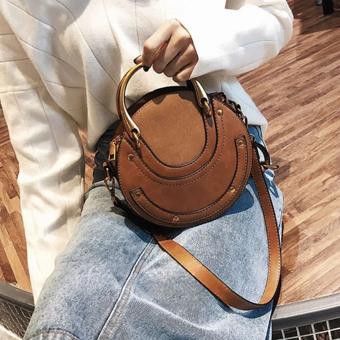 Woman holding round brown leather crossbody handbag, wearing white sweater and jeans