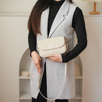 Woman wearing light gray vest and black top holding a beige crossbody bag indoors