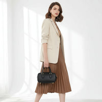 Woman in beige blazer and brown pleated skirt holding black leather handbag indoors