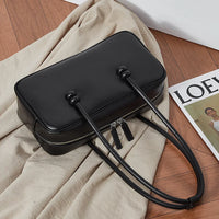 Black rectangular leather handbag with long handles on beige fabric and wooden floor