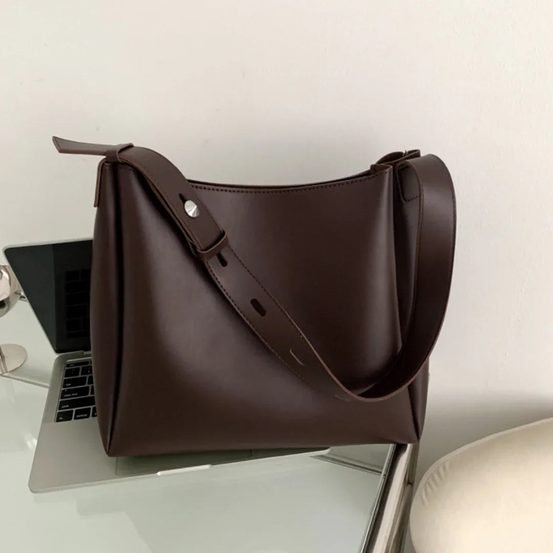 Minimalist brown leather shoulder bag on glass desk with laptop and lamp