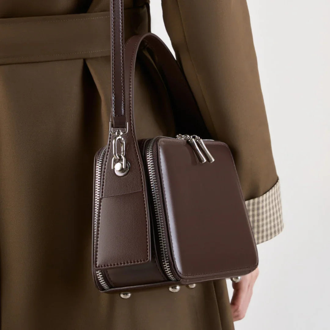 Brown square leather handbag with silver zippers and studs, worn with a brown coat