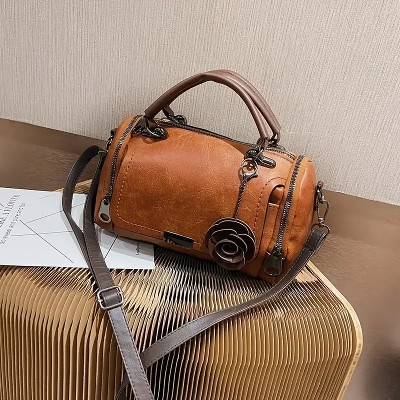 Brown leather handbag with flower charm and dual handles on a modern textured table