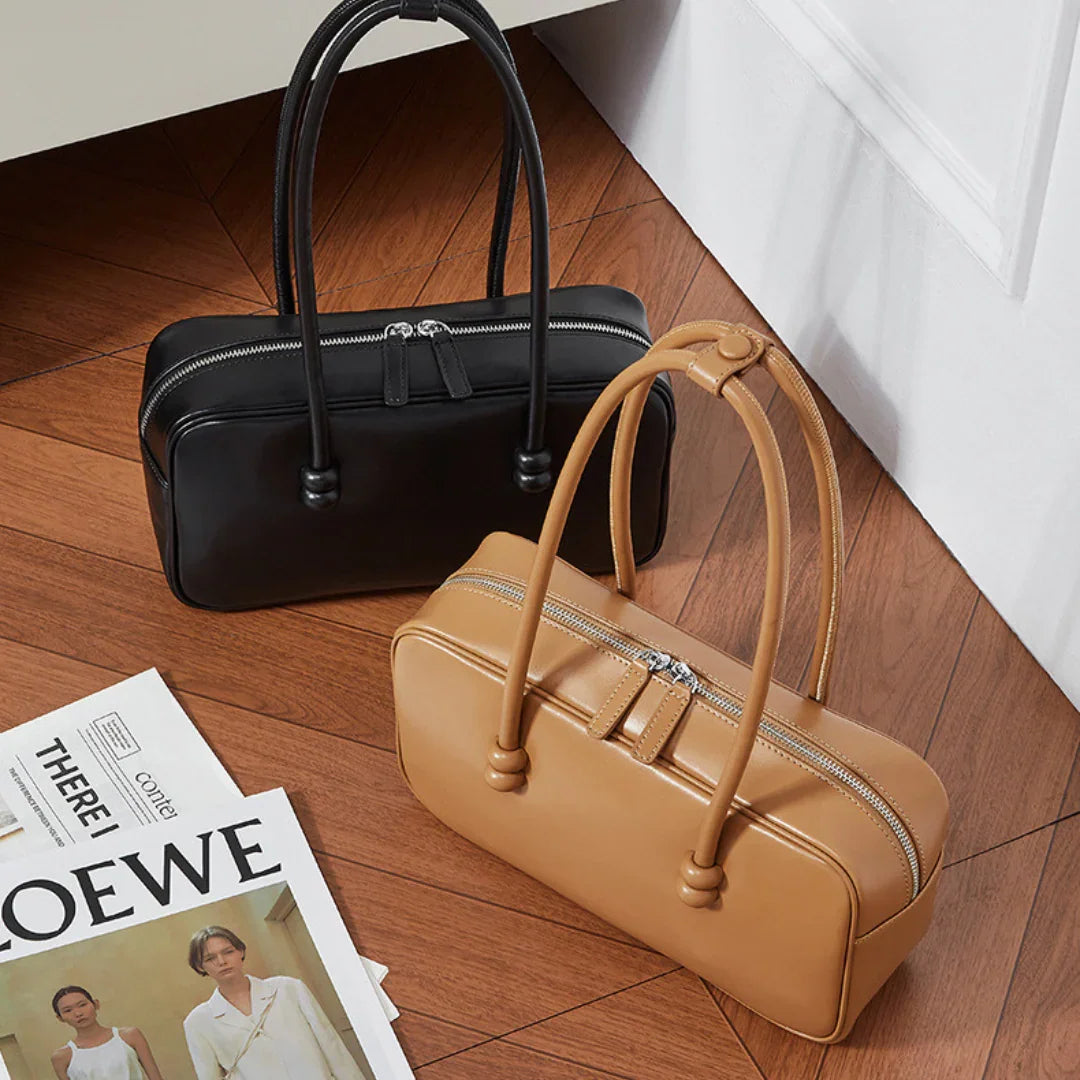 Black and brown leather handbags with top zippers on wooden floor next to magazines