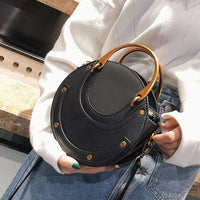 Round black handbag with gold handles, worn with white sweater and denim skirt.