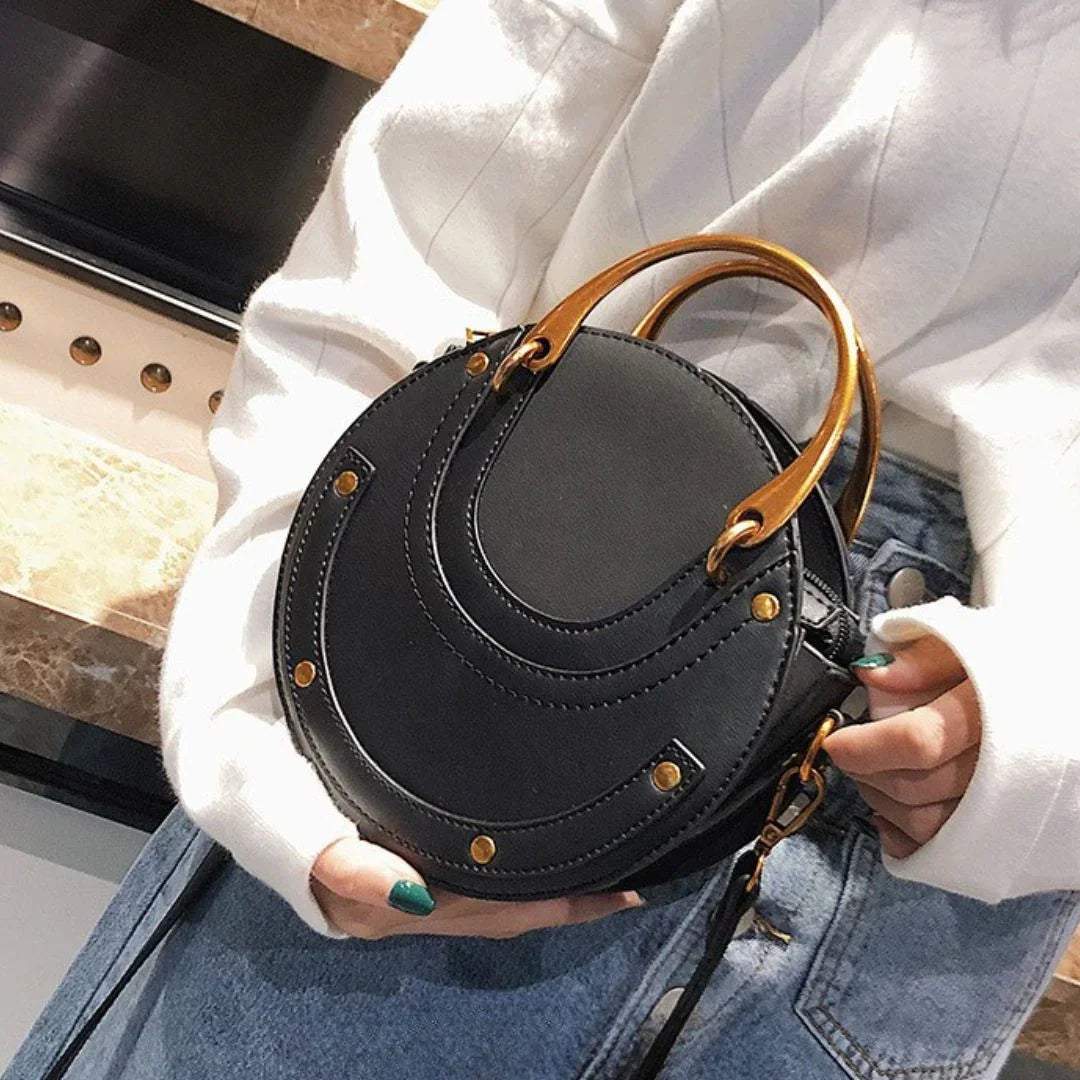 Round black handbag with gold handles, worn with white sweater and denim skirt.