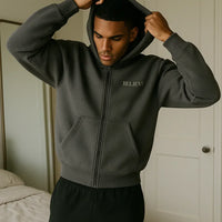 Man wearing a dark gray hoodie with 'BELIEVE' text, standing in a bedroom.