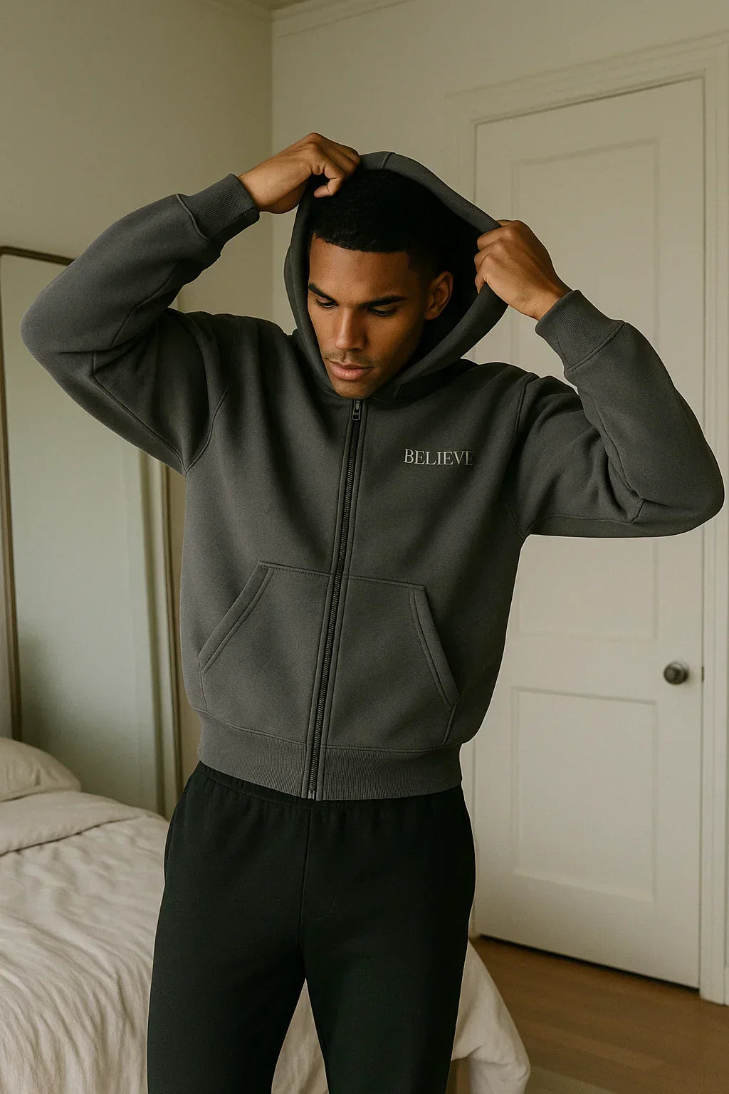 Man wearing a dark gray hoodie with 'BELIEVE' text, standing in a bedroom.