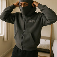 Person wearing a dark hoodie with 'BELIEVE' text in a bedroom setting
