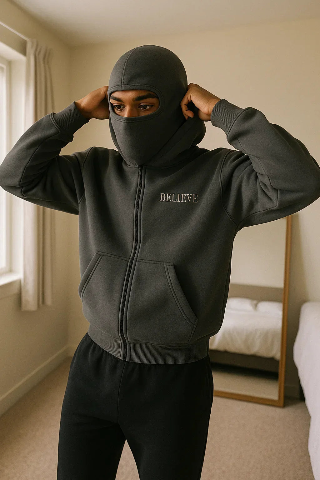 Person wearing a dark hoodie with 'BELIEVE' text in a bedroom setting