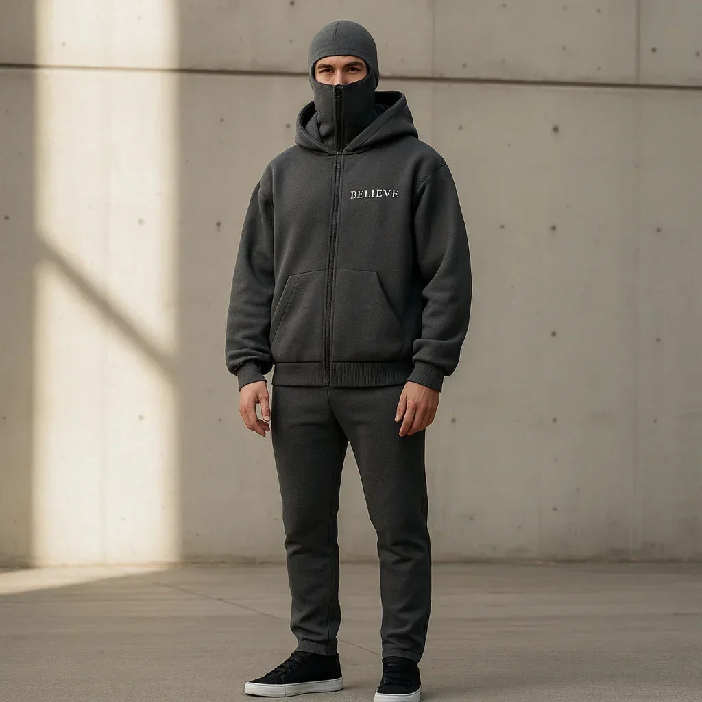 Person wearing a dark gray hoodie and pants with 'BELIEVE' text, standing against a concrete wall.