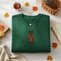 Green sweatshirt with a cat design and text, placed on a surface with autumn leaves and a cup of tea.