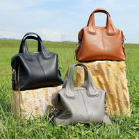 Three leather handbags in black, brown, and gray on stone blocks in a grassy outdoor setting