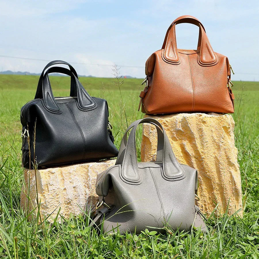 Three leather handbags in black, brown, and gray on stone blocks in a grassy outdoor setting