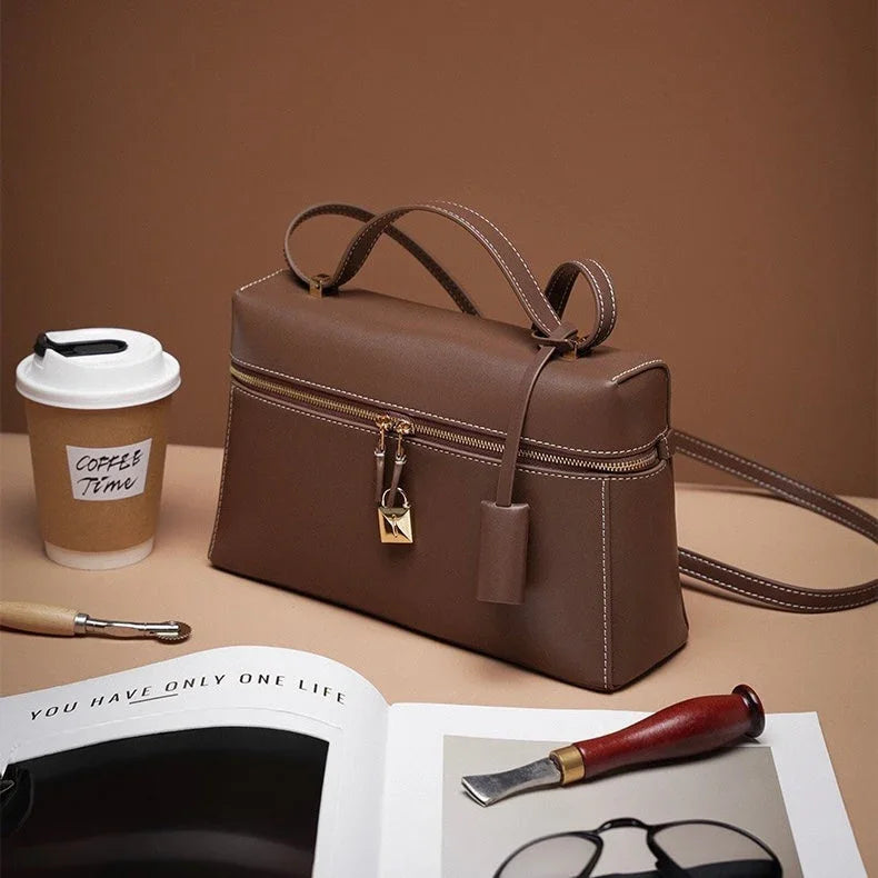 Brown leather handbag with gold zipper and lock, coffee cup, magazine, and carving tool on beige background