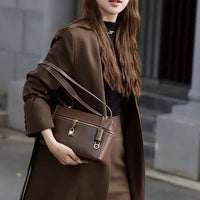 Woman in brown coat holding brown leather shoulder bag with gold accents outdoors