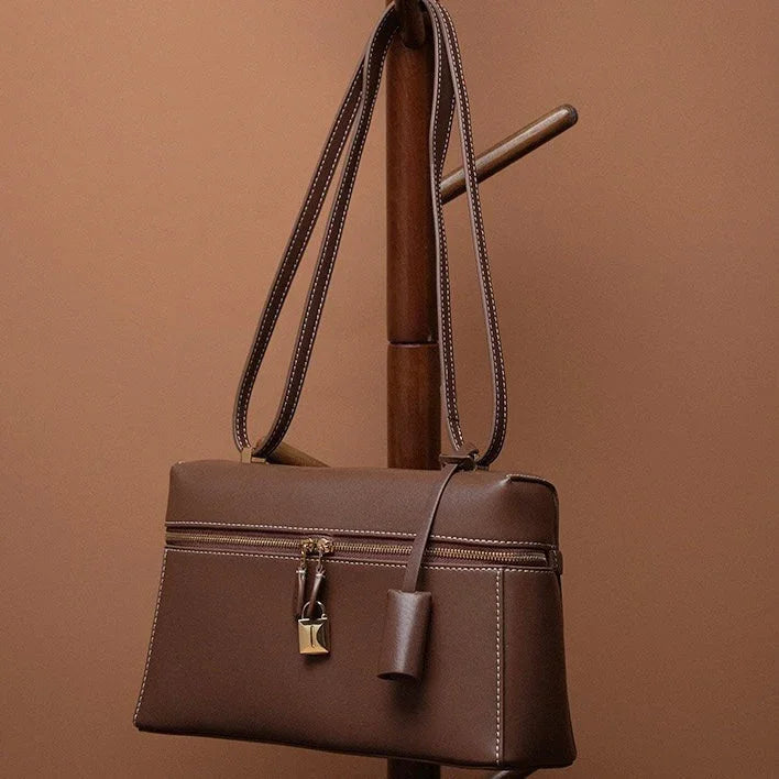 Brown leather handbag with lock detail on wooden stand against brown background