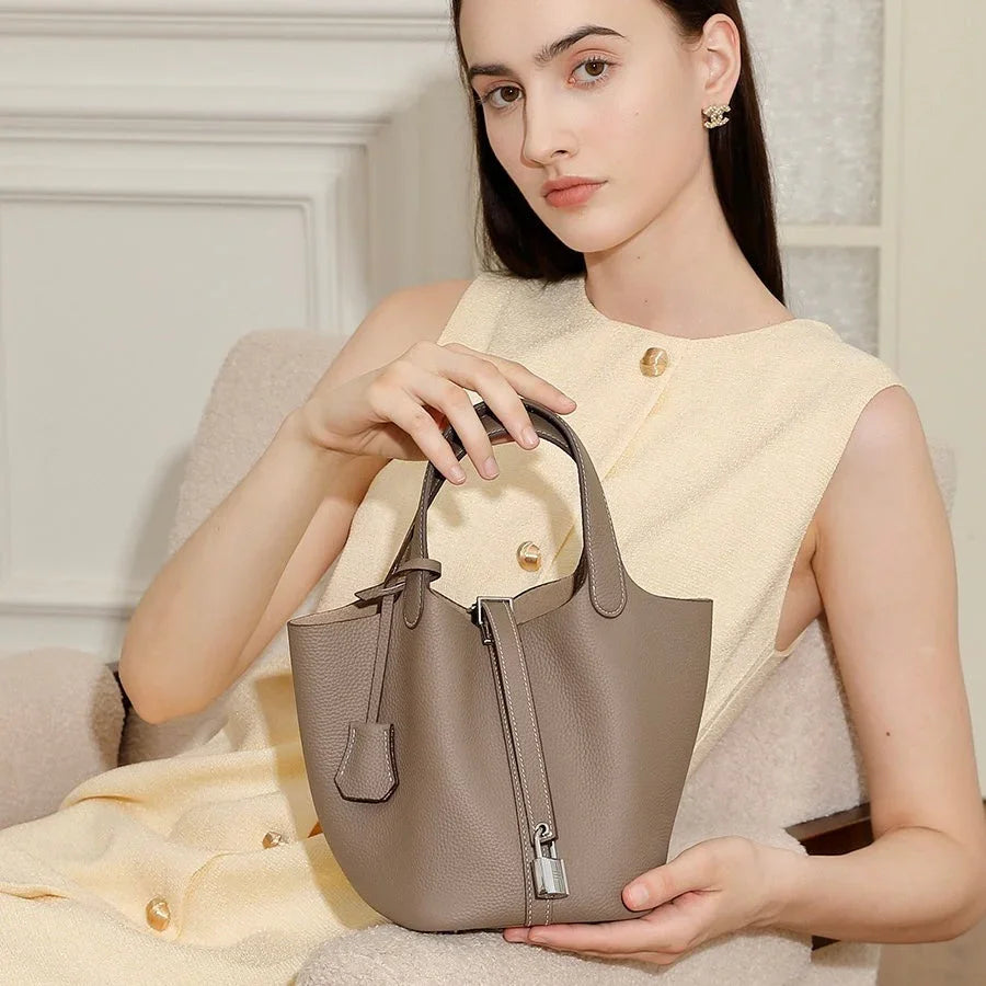 Woman in beige dress holding taupe leather handbag with silver lock, sitting indoors